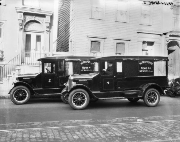 View across street towards two men driving two trucks. The signs painted on the side of the enclosed trucks reads: "Metropolitan News Co." Large buildings are in the background.
