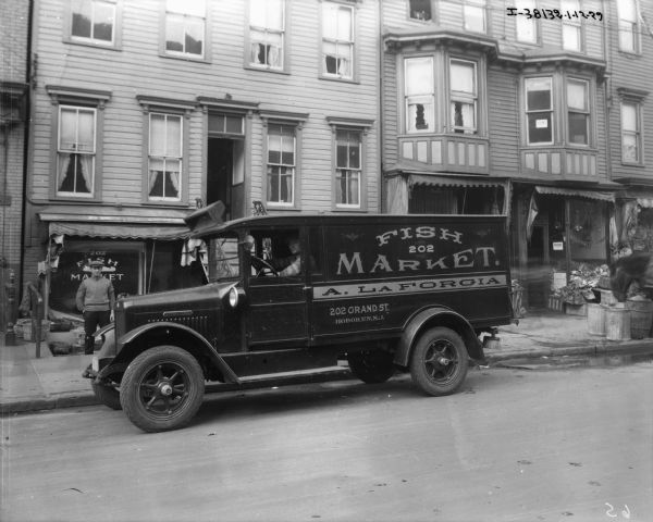 View across street towards a man sitting in a fish delivery truck. The sign on the side of the enclosed truck reads: "Fish Market, A. La Forgia, 202 Grant St. Hoboken NJ" A man is standing on the sidewalk behind the truck in front of a storefront that has a sign the reads "Fish Market." 