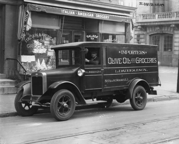 View across street towards a man sitting in the driver's seat of a delivery truck parked along the curb in front of an Italian-American Grocery. The sign on the truck reads: "Importers, Olive Oil and Groceries, L. Daddea & Son."