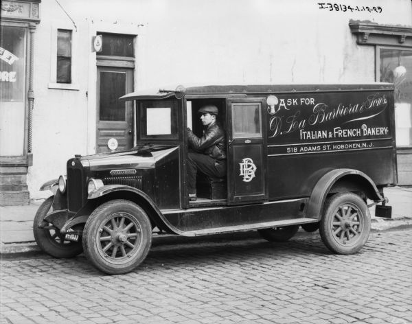 View across cobblestone street towards a man sitting in the driver's seat of a delivery truck parked along the curb in front of a building. The sign painted on the side of the enclosed truck reads: "D. La Barbiera & Sons, Italian & French Bakery 518 Adams St. Hoboken NJ"