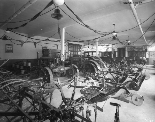 Agricultural machinery, tractors, and a truck on display on the floor of a dealership. Two men are sitting in chairs near a counter in the background.
