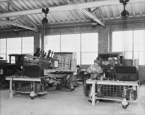 Men are working in a repair shop. There are large windows along the back wall in the background, and the ceiling is exposed. A block and tackle is hanging from a beam over the work areas on the left and right. One man is crouching on the floor and working at the rear axle of a truck in the center. The sign on the side of the stake body of the truck reads, in part: "State of Ohio, Dept. of Agriculture." Another man on the right is working at a worktable in front of a another truck.