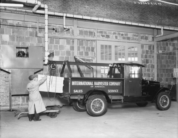 View across shop floor towards a man loading a crate onto an International Harvester Company of America sales and service truck. The man is using a Ford Triblock 1 ton block and tackle mounted on the truck. There are large doors or windows in the brick wall behind the truck.