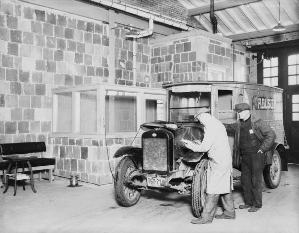 Man checking engine of a truck in a repair shop. The sign on the side of the truck reads: "Holsum Baking Co." The license plate on the front of the truck reads: "Ohio 1929." Against the wall on the left is a bench, table, and spittoon.