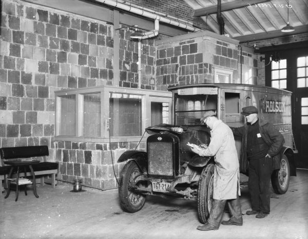 Man checking engine of a truck in a repair shop. The sign on the side of the truck reads: "Holsum Baking Co." The license plate on the front of the truck reads: "Ohio 1929." Against the wall on the left is a bench, table, and spittoon.