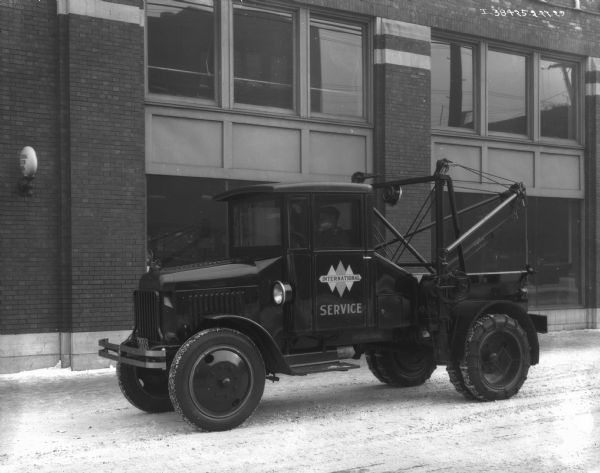 Driver's side view of a man driving an International Service truck with the triple diamond logo on the door. There are snow chains on the back wheels of the truck. Snow is on the ground, and an International Harvester building is in the background.