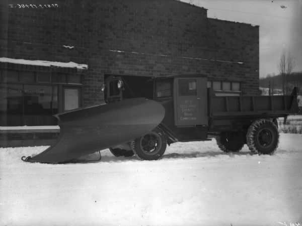 View towards driver's side of a snowplow parked in front of a brick building. The sign painted on the window reads: "[Leela]nau County Road Commission." Snow is on the ground.