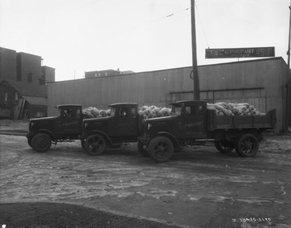 Three men are sitting in three trucks loaded with sacks in open truck beds parked in a row at an angle in Canada. The signs painted on each trucks driver's side door read: "C. Poupart, Contracteur." Industrial buildings are in the background, and on top of the building on the right is a large sign for "C. Poupart."