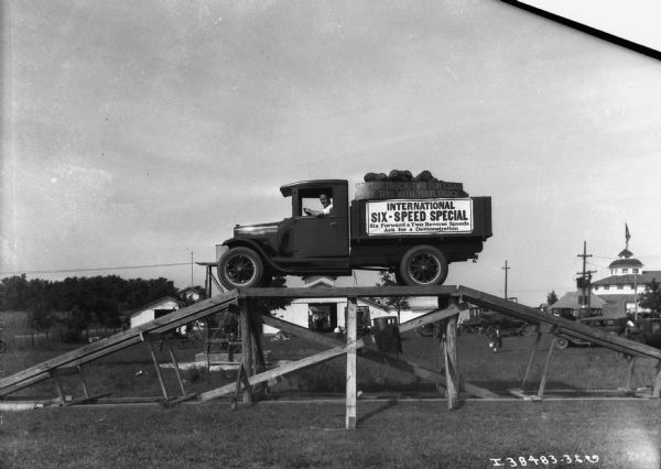 View towards a man driving a Six Speed special truck on top of a wooden ramp, on what may be fairgrounds. The sign along the side of the truck reads: "International Six-Speed Special, Six Forward & Two Reverse Speeds, Ask for a Demonstration." In the background trucks and automobiles are parked near barns and other outbuildings.