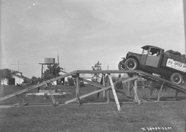 View towards a man driving a Six Speed special truck up to the top of a wooden ramp, on what may be fairgrounds. The sign along the side of the truck reads: "International Six-Speed Special, Six Forward & Two Reverse Speeds, Ask for a Demonstration." In the background trucks and automobiles are parked near barns and other outbuildings.