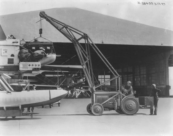 A man is driving a McCormick-Deering industrial tractor to work outdoors with what may be a seaplane. Another seaplane is under a hangar in the background. Men are working up on top of the seaplane, and other men are standing near the tractor.