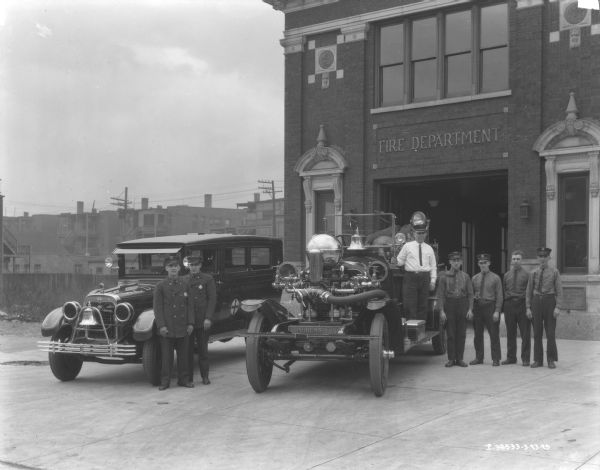 Group portrait of emergency ambulance personnel and fire fighters posing with an International ambulance and a fire truck in front of a fire station. The hood of the ambulance has an International logo on the front, and "C.F.D." is painted on top. The bell in front of the grill reads: "Fire Dept." On the front of the fire truck is a plate that reads: "Ahrens-Fox."