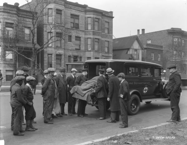 City of Chicago ambulance personnel are loading a person involved in an accident into an International ambulance as a small group of people are looking on. Painted on the side of the ambulance: "Fire Department C.F.D."