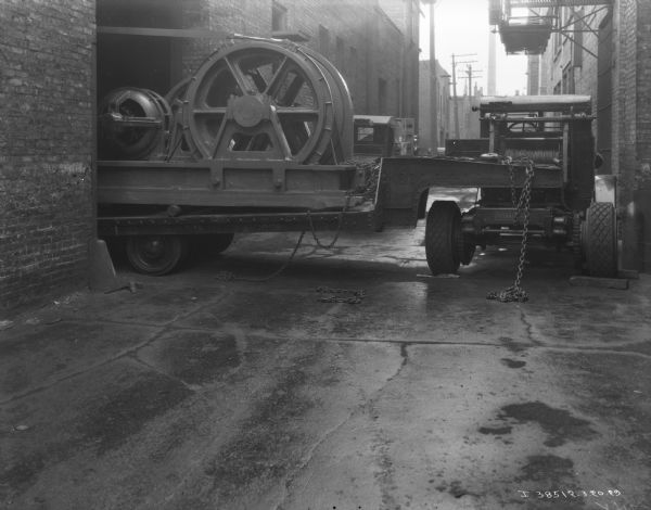 View down alley towards a truck with a tow cable mounted on it. A large section of machinery is on a trailer attached to the truck, and the trailer is halfway inside a large opening in a brick building.