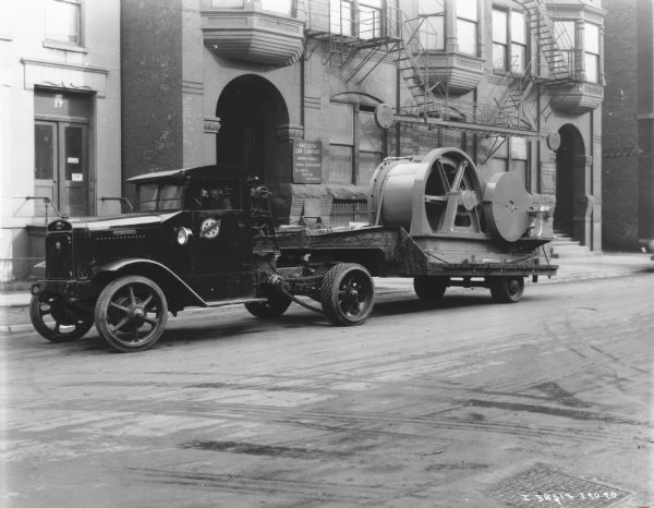 View towards a man sitting in the driver's seat of a truck with a tow cable. A sign on the driver's side door reads: "Thomas Service." The truck is hauling a large piece of machinery on a trailer. A sign on the building in the background reads: "Vacuum Can Company."