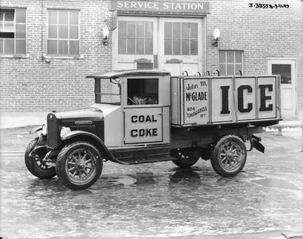 View of driver's side of delivery truck. Signs painted on the truck read: "Coal, Coke," and John M. McGlade, 874 Tonawanda St., ICE." A brick building behind the truck has a sign above the garage doors that reads: "Service Station."