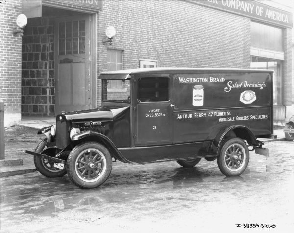 View of the driver's side of a delivery truck. The signs painted on the side of the truck read: "Washington Brand Salad Dressings, Arthur Ferry 47 Flower St., Wholesale Grocers Specialties." A large brick building in the background has open garage doors, and a sign for "[International Harvester] Company of America."