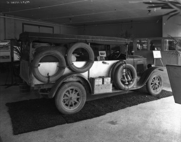 Passenger side view of the "Truck That Crossed The Sahara." The truck is parked indoors at what may be a dealership. Spare tires are tied to the side of the truck, along with glass lanterns, and cans of No. 1 Sphinx in a box on the running board. A display rack in the background on the left has a sign for "Sahara" and photographs. An office is in the background on the right.