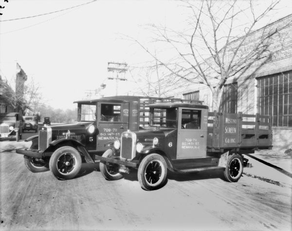 View towards two delivery trucks parked at an angle in the street. On the right is a large brick industrial building. The signs on the truck read: "Newark, New Jersey" and "Westnu[...] Screen Co. Inc."