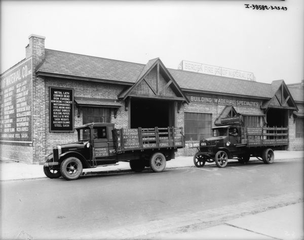 View across street towards two trucks parked n front of a large brick building with a sign on the roof that reads: "Bergen Fire Proof Material Co." A man is sitting in the driver's seat of the truck on the right. Signs on the trucks read: "Corner Bead, Steel Sash, Iron Furring, Metal Lath, Asphalt, Shingles, Bars."
