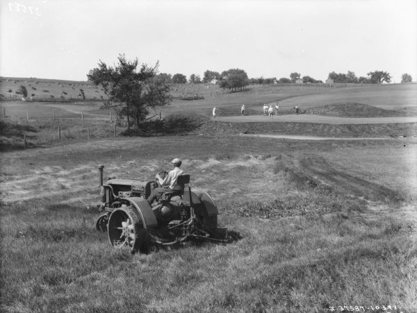 A man is using a Farmall tractor to pull a mower in the foreground. A group of men are golfing in the background. A field with harvested grain is on the hill to the left along the golf course.