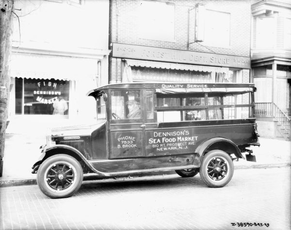 A man is sitting in the driver's seat of a delivery truck for  Dennison's Food Market which is parked along the curb in front of the market. Next to the market on the right is the Forest Hill Dept. Store.