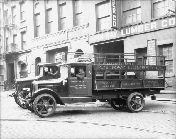 Two men are sitting in the driver's seats of two delivery trucks parked in front of the Pin-Ray Lumber Co. building. The sign on the driver side door of the truck in front reads: "13 Sheriff St. N.Y.C."