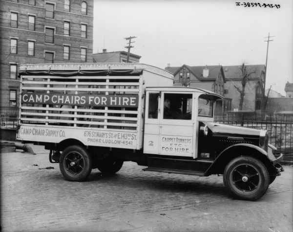 Two men are sitting in the cab of a truck parked outdoors. The signs on the side of the truck read: "Carpet Runners, Etc. For Hire," "Tables, Card Tables, Coat Racks & Canopies" and "Camp Chair Supply Co."