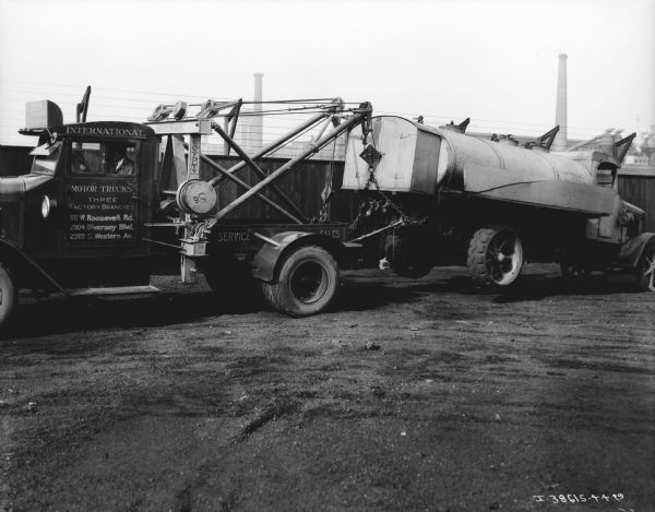 International Harvester Service Truck towing a truck. A man is sitting in the driver's seat of the truck. The sign painted on the truck reads, in part: "International Motor Trucks, Three Factory Branches."