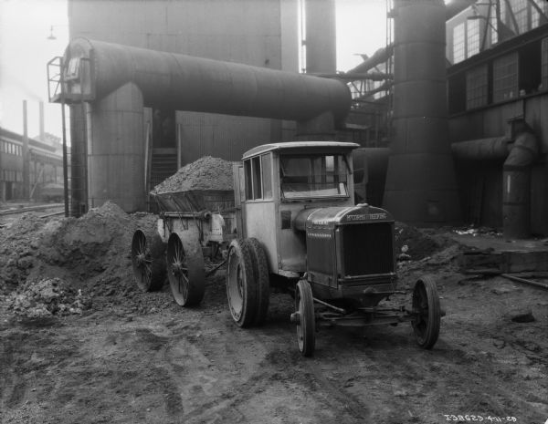 View towards the front of a McCormick-Deering tractor with an enclosed cab. The tractor is pulling a fully loaded trailer. In the background are factory buildings.