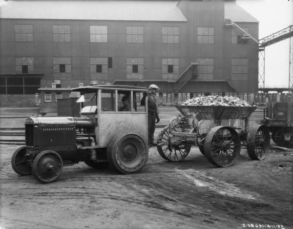 View towards a man driving an industrial tractor on the left, pulling a load of coal. Another man is standing on the trailer hitch behind the tractor. A factory building is in the background.