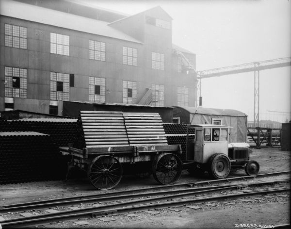 View towards the right side of an industrial tractor pulling a trailer with a load of pipe stacked inside. There is a factory building in he background. Railroad tracks are in the foreground.