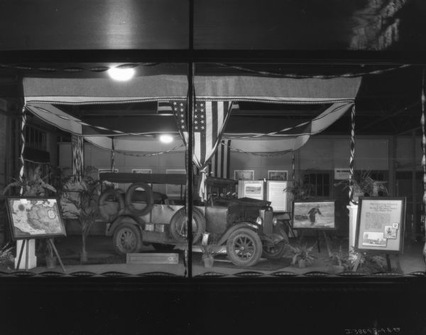 View through a show window of a display of the truck that crossed the Sahara. There is a map on an easel on the left. A poster on an easel on the right has a title that reads: "The First Four-Wheeled Stock Truck to Cross the Sahara Desert on Display Here."