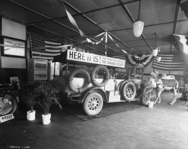 View of a display of the truck that crossed the Sahara. There is camel on the far right near the front of the truck. Banners and flags are hanging around the room. A sign above the trucks reads: "Here It Is! The Truck That Crossed The Sahara Desert."