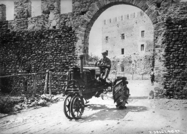 View towards a man who has driven a Farmall tractor through a large stone archway in a tall, stone wall. A large brick building is in the background.