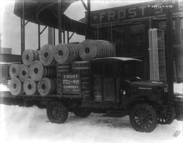 View towards passenger side of a delivery truck parked outdoors in the snow. The sign on the truck reads: "Frost Steel & Wire Company. Limited." There is a large industrial building in the background with a sign painted along the top that reads in part: "Frost."