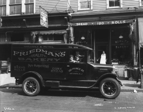 View across street towards a delivery truck parked along the curb in front of Friedman's Bakery at 120 Prince Street. Two children are sitting in the passenger seat of the truck looking out of the window. A man is sitting in the driver's seat, and a woman is standing at the door of the bakery.