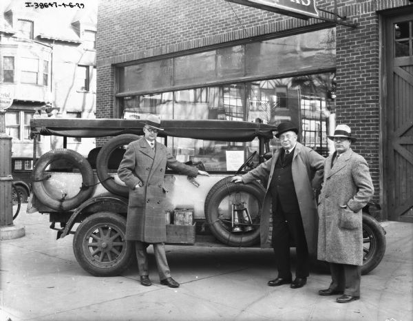 Three men are standing by the "Truck That Crossed Sahara Desert" on display outdoors. In the background is a brick building with large glass windows. There is a gas pump on the far left.