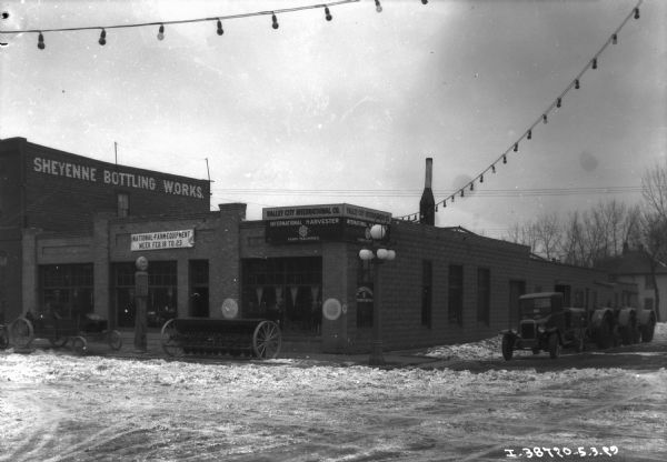 View from street towards a dealership with a sign that reads: "Farm Equipment Week Feb. 18 to 23." On the building next door on the right is a sign that reads: "Sheyenne Bottling Works."