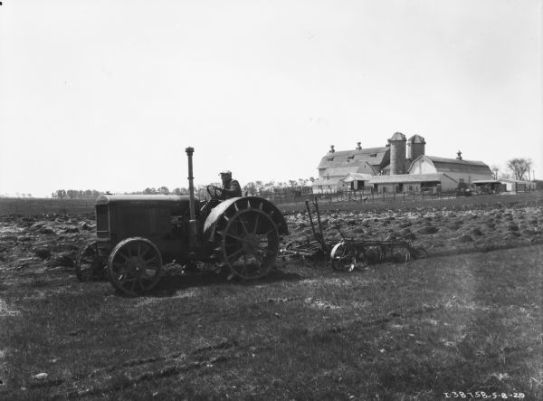 View towards a man using a tractor to pull an agricultural implement in a field. There are barns and silos in the background.