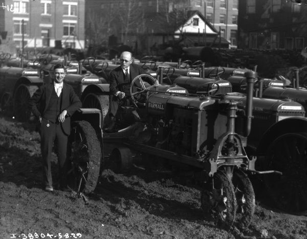 Two men are posing with a large lot full of Farmall tractors. There are buildings in the background.