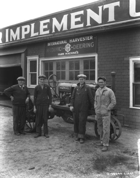 Men are posing with a Farmall tractor in front of a building with a sign for IH and McCormick-Deering farm machines.