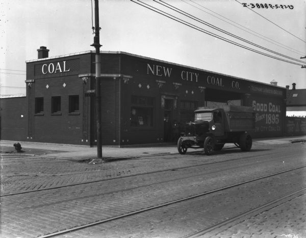 View across road towards a man sitting in the driver's seat of a truck parked in front of a building with a sign that reads: "New City Coal Co." A group of men are standing at the entrance.