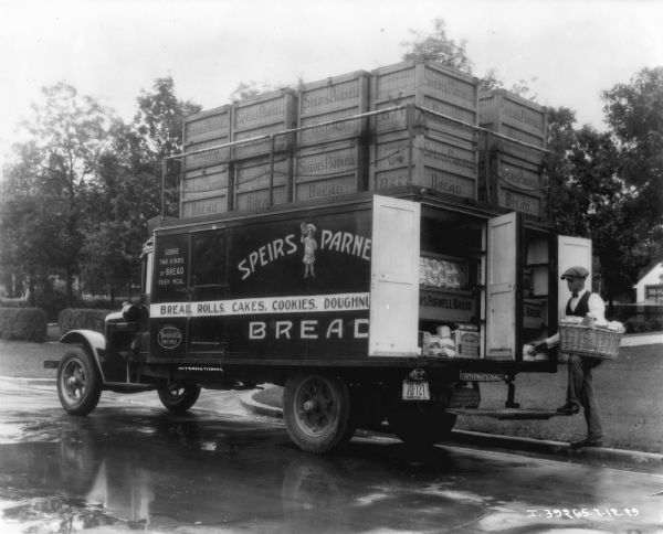 Three-quarter view from left rear of a delivery truck parked at a curb. A man is holding a basket while standing at the back of the truck, which has it's back doors open. There are large crates staked on the top of the truck secured behind a metal railing and chains. The sign on the side of the truck reads: "Speirs Parnell Bread." Manitoba, Canada.