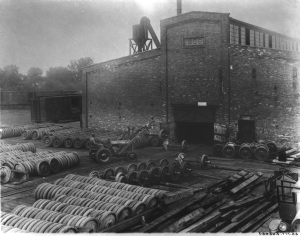 Elevated view of a railroad yard. A large, brick building has a date stone of "1908" and railroad tracks and railroad cars are along the building on the left. Wheels and wheels with axles are lined up in front of the building, and a man is using an industrial tractor and a winch to load the railroad cars.
