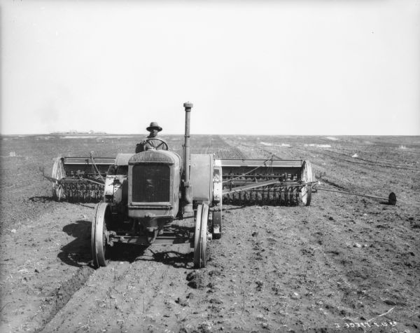 Front view of a man driving a McCormick-Deering tractor pulling a disk harrow with power lift. There are buildings in the far distance on the left.