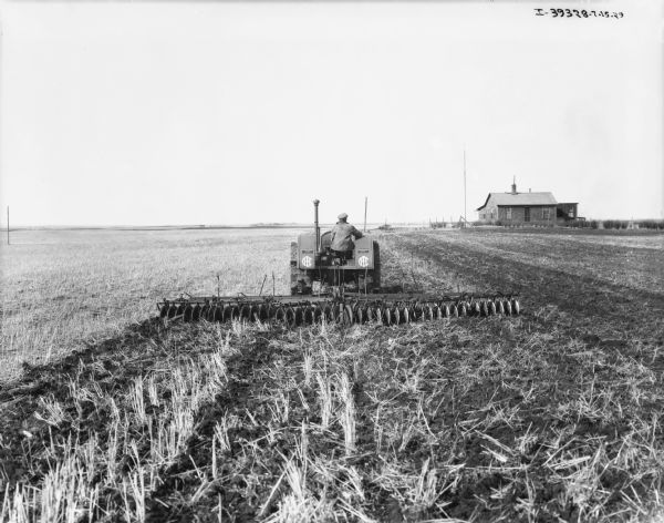 Rear view of a man driving a tractor pulling a disk harrow in a field. Farm buildings are in the background on the right.
