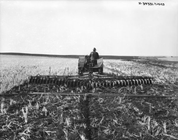Rear view of a man driving a tractor pulling a disk harrow in a field.