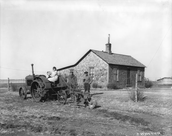 Family of three people posing with tractor pulling a disk harrow in front of a small house.