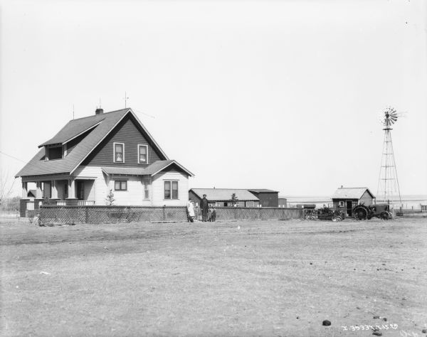 A man and women and two children are posing in front of a fence surrounding a small house. There is a tractor, an automobile, agricultural implements, and a windmill on the right.